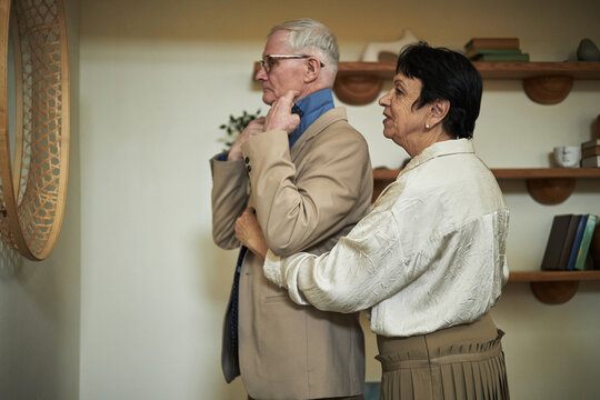 Senior woman assisting elderly Caucasian husband with choosing right tie, man getting dressed in front of mirror for going out, couple chatting - Powered by Adobe