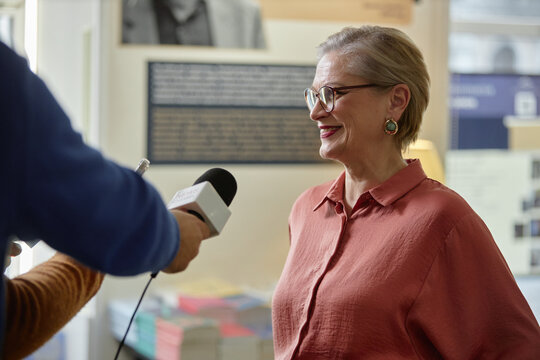 Caucasian middle aged woman smiling while being interviewed by unseen holding microphone, standing indoors with blurred books and informational displays in background