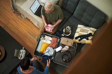Overhead view of elderly couple diligently packing suitcase on couch with various items carefully...