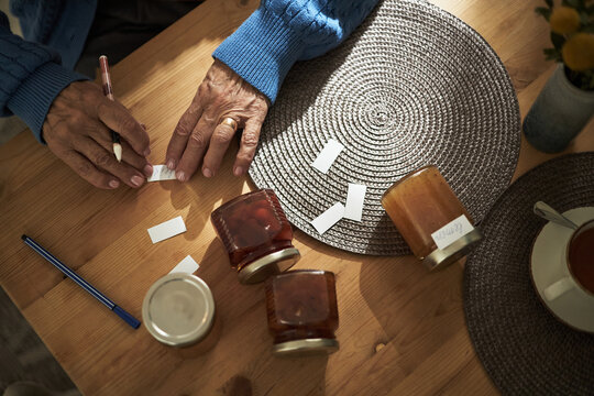 Senior woman labeling jars of homemade jam at wooden table with pen and paper labels scattered around. Cozy and relaxed atmosphere during preparation for market or gift-giving
