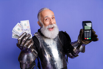 Elderly man in knight costume holding money and card reader against purple background