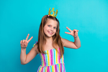 Cute girl in a colorful striped dress wearing a golden crown shows peace gestures on a bright blue...