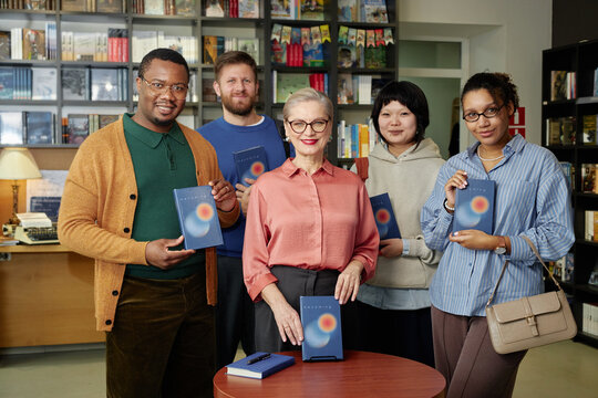 Group of diverse adults posing with senior Caucasian female famous author standing together in bookstore holding books, smiling at camera after autograph session