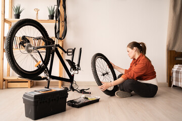 A young female bicycle mechanic sits on the floor, carefully examining the front wheel of her...