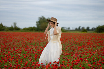 Young woman in a white dress and hat walks among summer poppies