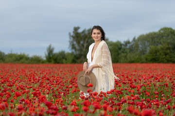 Young woman in a white dress and hat posing among summer poppies