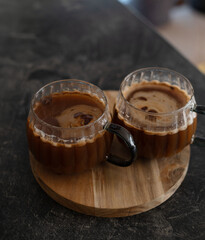 two glass mugs filled with rich dark coffee placed on a wooden tray, resting on a dark textured surface under soft ambient lighting in a cozy indoor setting, delicate, stylish, classic, fashion