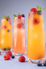 three tall glasses filled with refreshing orange and pink beverages garnished with fresh raspberries, blueberries, and mint leaves, set against a cool-toned background conveying a vibrant, summery vib