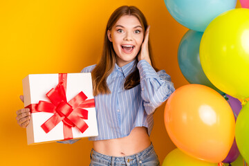 Cheerful young woman holding a gift box with a red ribbon, surrounded by vibrant balloons on a yellow background