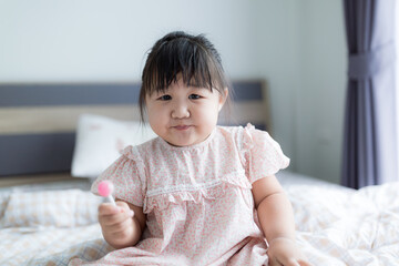 Fototapeta premium Cute little girl is happily eating a lollipop. Asian sweet tooth girl is holding a small lollipop sitting on the bed at home.