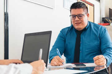 A man in a blue shirt and black tie is writing on a piece of paper