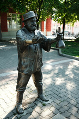 Full-body bronze sculpture of a man holding grapes and a wine bottle, standing in a paved park area in Vrsac, Serbia country.
