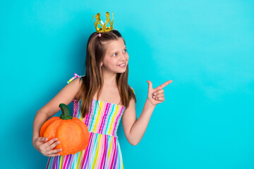 Charming young girl with a pumpkin in colorful dress posing playfully on a vibrant teal background,...