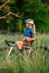 Attractive blonde woman in cycling gear and helmet on gravel bike, standing in tall grass. Concept of active lifestyle, fitness, outdoor adventure, and freedom.