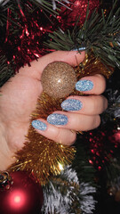 Hand with sparkly blue nails holding golden ornament, decorated with festive garland