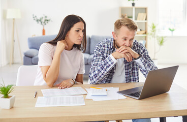 Young worried couple sitting at the table with laptop and calculating finances or taxes at home....