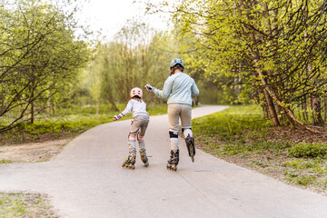 Two girls rollerblading on park path in spring, wearing helmets and knee pads. Children enjoy...