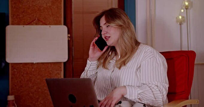 A focused young woman handles remote work from home, engaging in a phone call and typing on her laptop. She also reviews notes in a red notebook, showing her multitasking skills.
