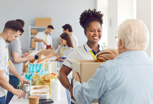 Happy smiling african american volunteer working at charity center giving free food donations for mature man in charitable foundation. Humanitarian aid, volunteering and social help for poor concept.
