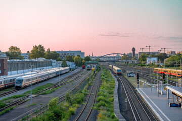 Trains in the depot and on the tracks under a pink sky. An old water tower and modern buildings dominate the horizon.