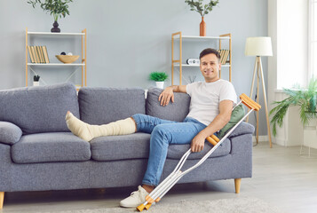 Young smiling man with crutches sitting on sofa at home on rehabilitation with leg in plaster cast...