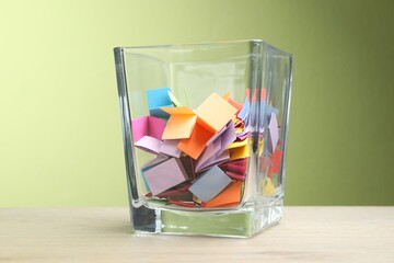 Jar with colorful pieces of paper on wooden table against green background, closeup