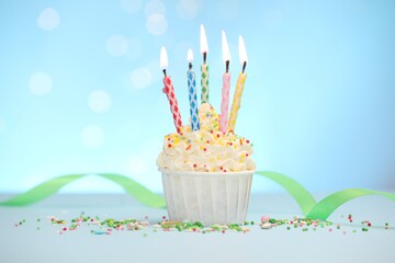 Burning candles in cupcake and sprinkles on white table against light blue background with blurred...