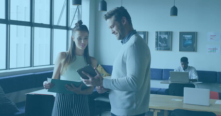 Two colleagues discussing documents and tablet in office lounge, with wooden tables