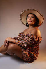 Gorgeous brown skin woman wearing a straw hat and floral pattern dress, sitting on the ground. Perfect for vacations, beauty, summer, lifestyle. Studio portrait with copy space.