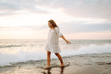 Happy young woman in white dress enjoying sunset rays on seashore. Beautiful woman walking along...