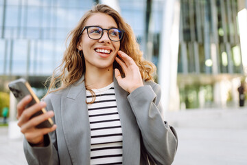 Young woman in stylish clothes and holding phone enjoying sunny day near modern building. Stylish woman looking at smartphone and smiling outdoors. Enjoyment and relaxation concept. Lifestyle.