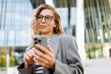 Young woman in stylish clothes and holding phone enjoying sunny day near modern building. Stylish...