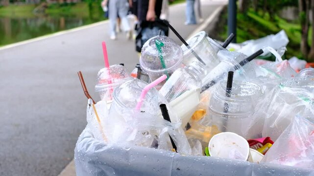 A trash can full of plastic cups and straws. The trash can is on the side of a road