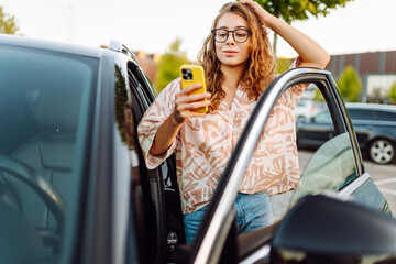 Naklejka premium Beautiful woman with curly hair with a phone in her hands stands near a car in a parking lot. Young female driver enjoys the weather and chats at sunset. Transport and technology concept.