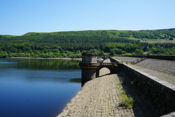 Fototapeta premium Derbyshire, England - June 20 2025: Scenic view of a reservoir with a dam structure and tower structures reflecting the forest and hills under a clear blue sky.