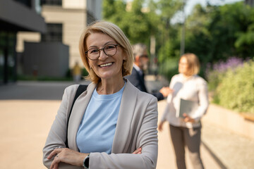 Beautiful mature businesswoman with crossed arms smiling confidently outdoors in urban business district, looking at camera

