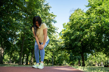 Beautiful woman experiencing knee pain while running on trail in park during morning, outdoor...