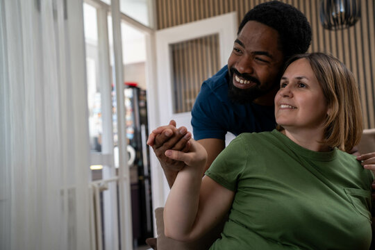 Happy interracial couple holding hands looking together away in bright living room, positive mood and warm daytime atmosphere