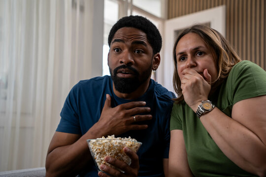 Tense interracial couple watching thriller horror movie with popcorn in bright living room during daytime

