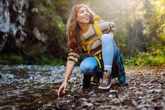 Beautiful woman in hiker with bright yellow backpack on travel touching mountain river with hands and enjoying journey. Young female traveler on hiking trail in mountains. Active lifestyle.