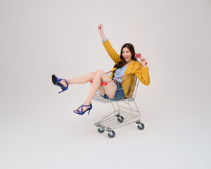 Ecstatic Asian woman in yellow blazer and heels holding a credit card joyfully sits in a shopping cart. Perfect for sales, promotions, or payment themes.