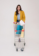 Joyful Asian woman in yellow blazer pushes a shopping cart filled with shopping bags isolated on white background.