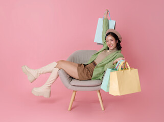 Joyful Asian woman in stylish outfit with beret relaxes in a chair holding up colorful shopping bags isolated on pink background