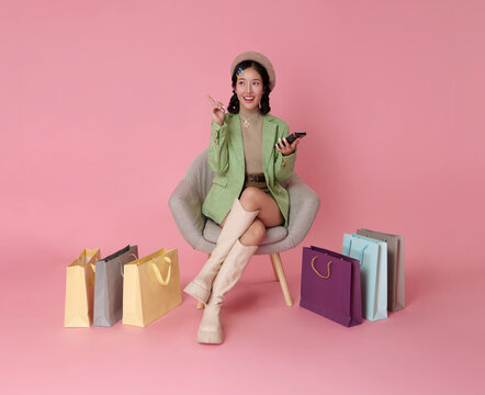 Joyful Asian woman in stylish outfit with beret relaxes in a chair holding mobile phone in hands shopping online isolated on pink background. - Powered by Adobe