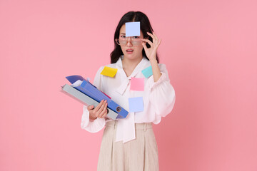 Asian woman feeling stressed with work overload, covered in colorful sticky notes and holding folders. Studio portrait on pink background, representing office pressure and multitasking.