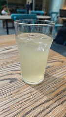 Glass of cold lemonade with ice cubes on a wooden table in a cozy cafe setting. The image evokes refreshment, summer relaxation, and casual social moments