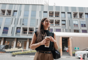 Young Asian businesswoman holding a takeaway coffee walking in the city and looking away