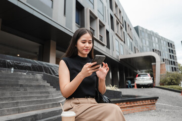 Businesswoman using mobile phone app sitting on steps in front of modern office building with takeaway coffee