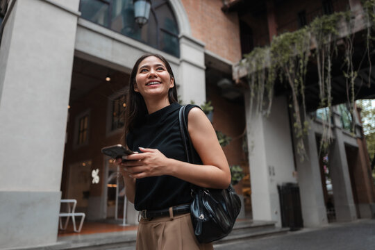 Happy businesswoman holding mobile phone and smiling while walking in city street
