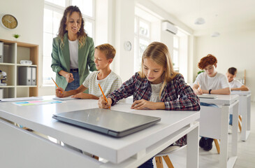 Obraz premium Female friendly woman teacher helping a boy during a lesson in the classroom in junior high school. Schoolchildren sitting at the desks and working. Education, knowledge and back to school concept.